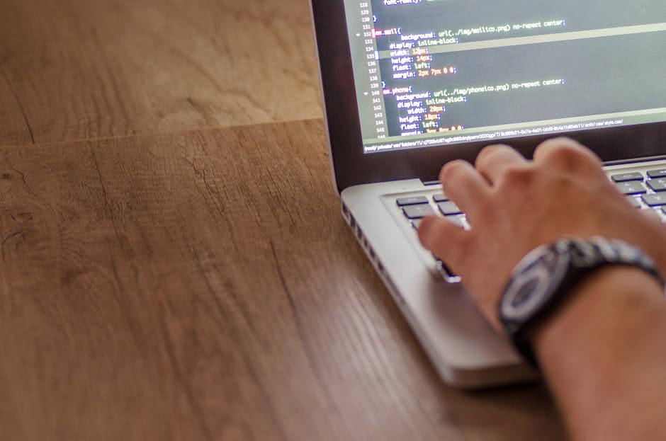 A close-up of a person's hand typing on a laptop keyboard placed on a wooden desk, with visible lines of code on the screen. The person's wrist is adorned with a watch with a dark strap. The backgroun