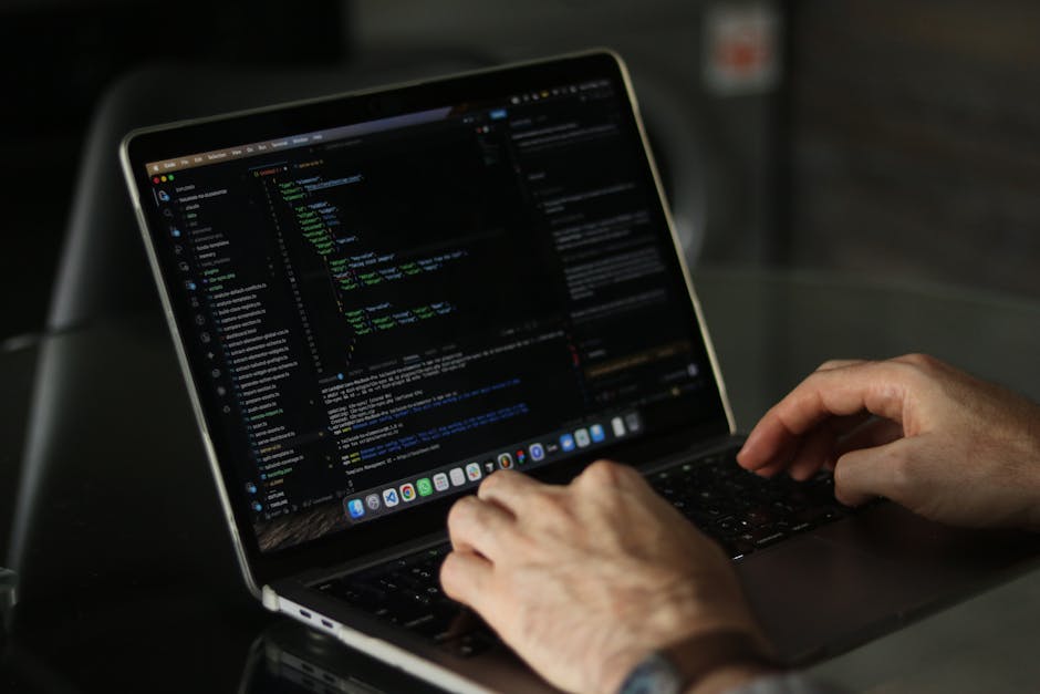 A person's hands are seen typing on a laptop with a dark screen displaying lines of code in different colors, suggestive of programming or web development work. The laptop, positioned on a reflective 