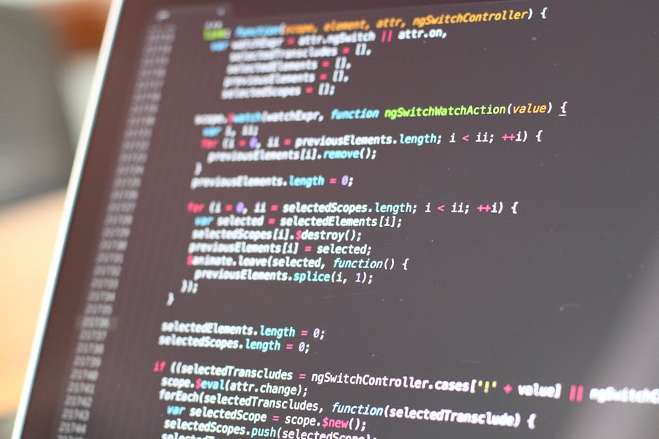A person's hands are seen typing on a laptop with a dark screen displaying lines of code in different colors, suggestive of programming or web development work. The laptop, positioned on a reflective 